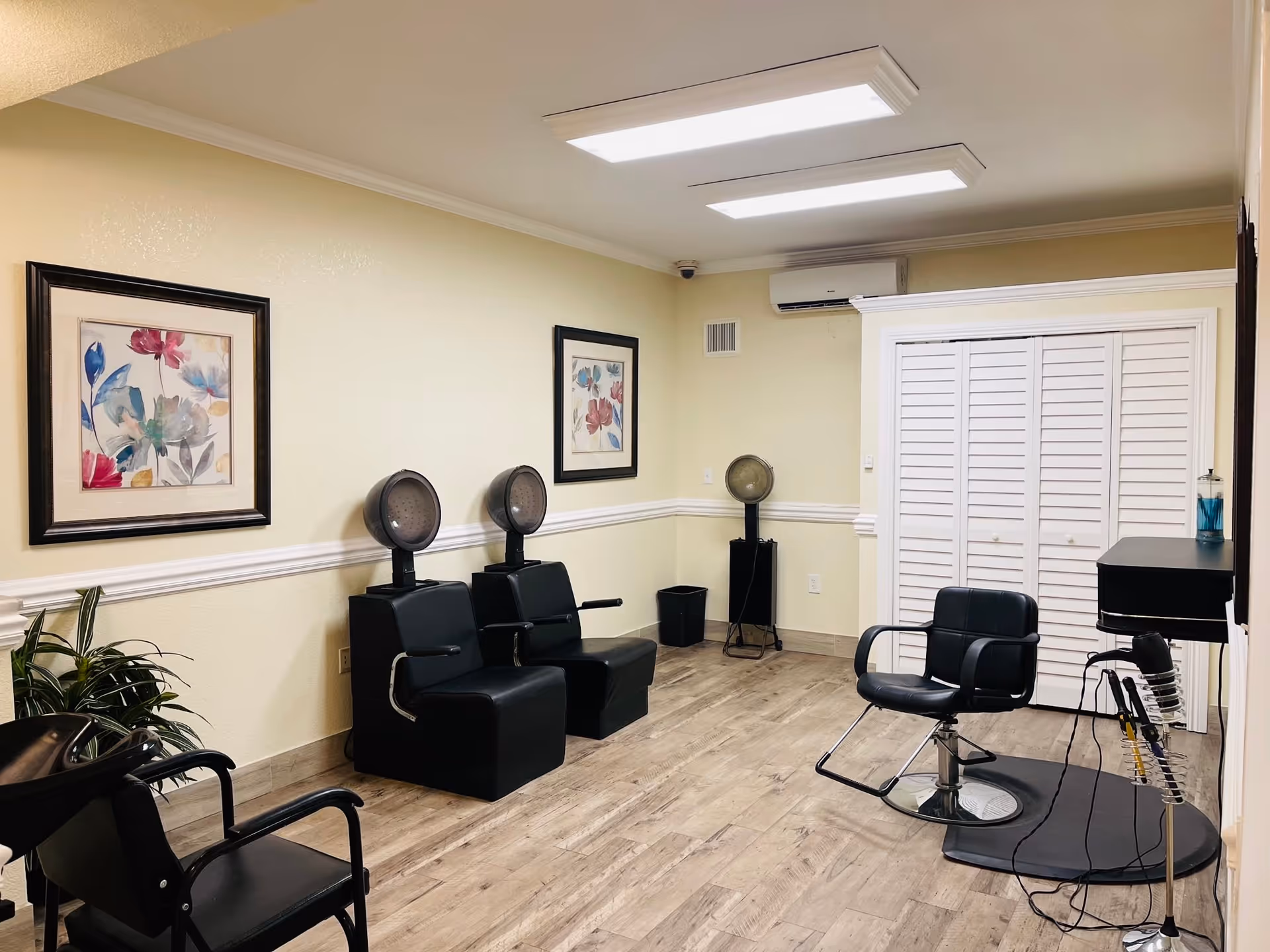 Interior of a hair salon area with two black hair drying chairs, a black styling chair in front of a mirror, wooden flooring, two framed floral paintings on the wall, and a white louvered door in the background.
