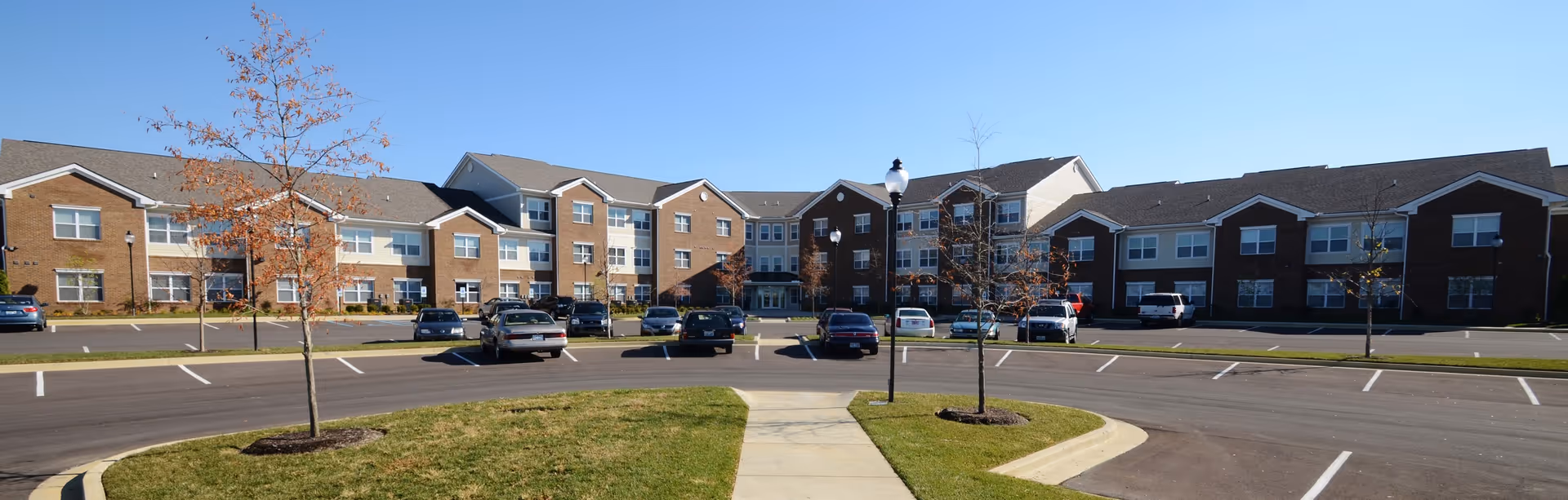 Wide exterior view of Hamburg Senior Residence, a large multi-story senior living building with brick and beige siding. There is a parking lot with several cars parked and a sidewalk leading to the main entrance. Small trees and grass areas are visible in the foreground under a clear blue sky.