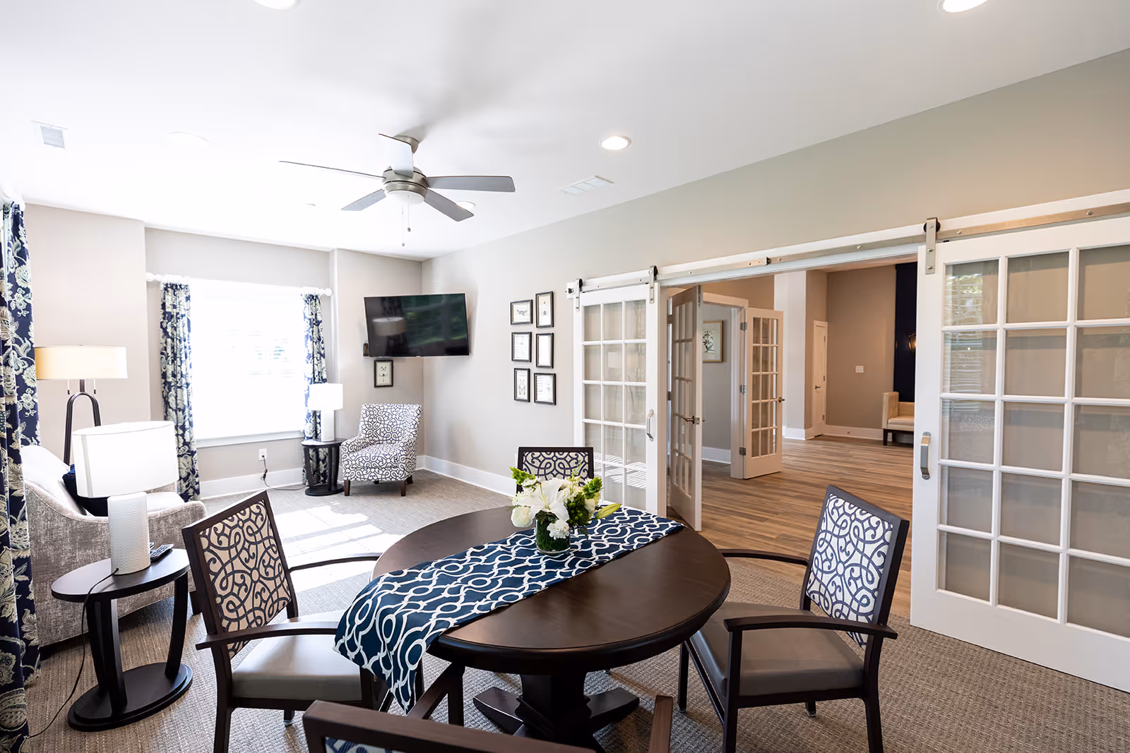 A bright and airy living room area with a round wooden table covered by a blue and white patterned runner, surrounded by four chairs with matching patterned cushions. The room features large windows with blue and white floral curtains, a ceiling fan, a wall-mounted flat-screen TV, and sliding glass doors leading to another room with wooden flooring.