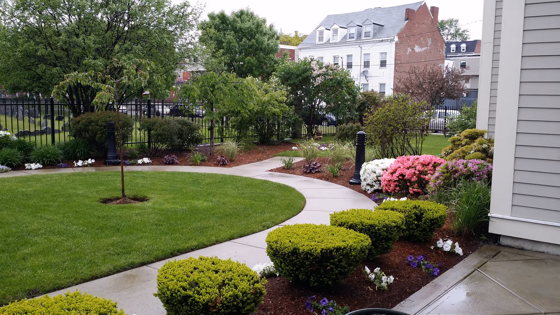 A well-maintained garden area with a curved concrete pathway surrounded by green grass, neatly trimmed bushes, and colorful flowering plants. Trees and shrubs line the pathway, and a black metal fence separates the garden from neighboring buildings in the background.
