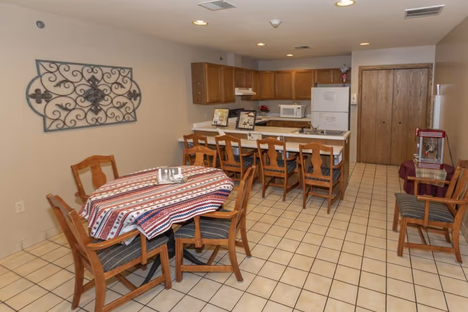 A dining area with a rectangular table covered by a patterned tablecloth and surrounded by six wooden chairs with striped cushions. Behind the table is a kitchen with wooden cabinets, a white refrigerator, microwave, and a counter with five wooden chairs. The floor is tiled, and there is a decorative wrought iron wall hanging on the left wall.