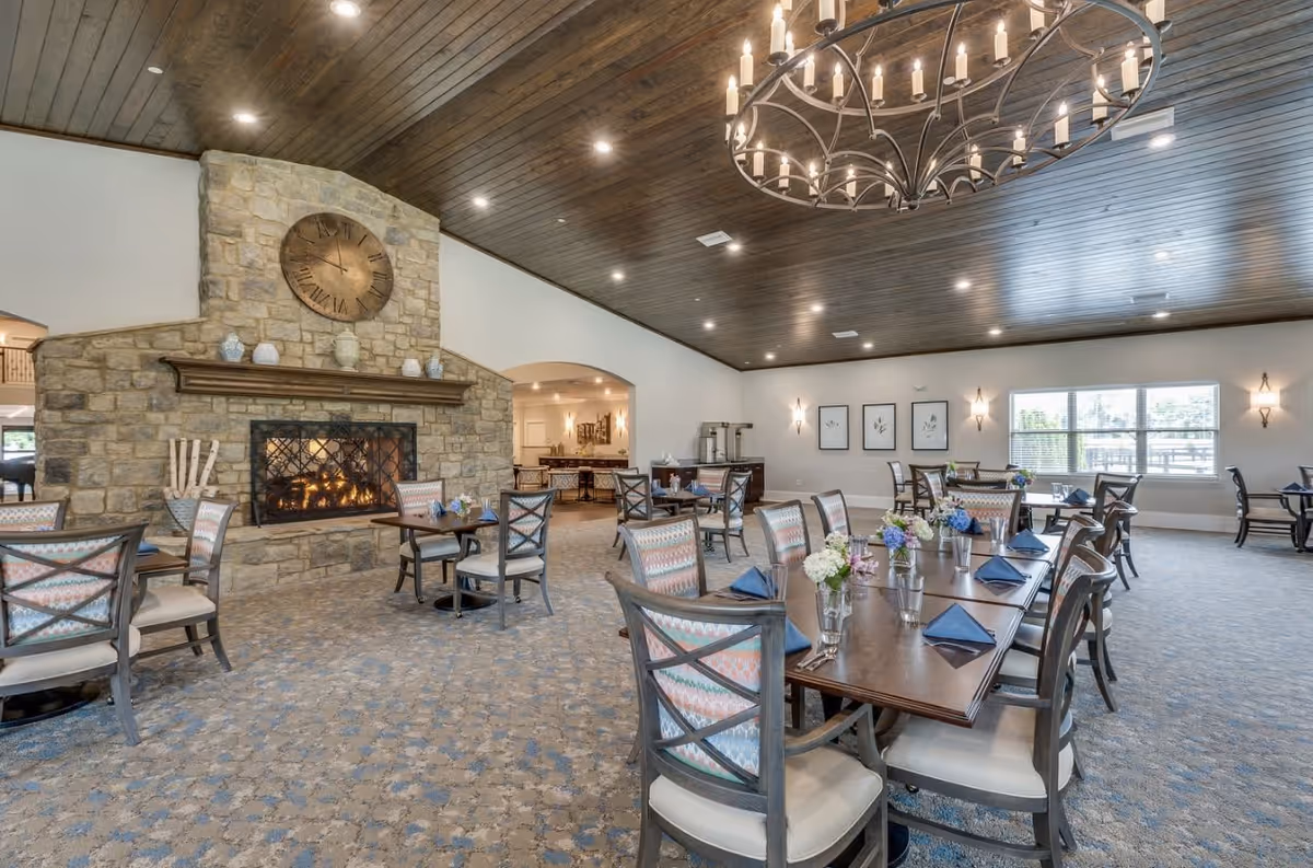A spacious dining room with multiple tables and chairs arranged neatly. Each table is set with blue napkins, glassware, and small floral centerpieces. The room features a stone fireplace with a large clock above it, a wooden ceiling with recessed lighting, and a large chandelier. There are windows letting in natural light and framed artwork on the walls.