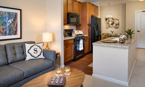Interior view of a senior living facility apartment showing a cozy living room with a gray couch, a decorative pillow with the letter 'S', a wooden coffee table with two glasses and a game, a lamp on a side table, and a colorful abstract painting on the wall. Adjacent to the living room is a kitchen area with wooden cabinets, a black refrigerator, a stove with a microwave above it, and a granite countertop island with place settings and decorative items.