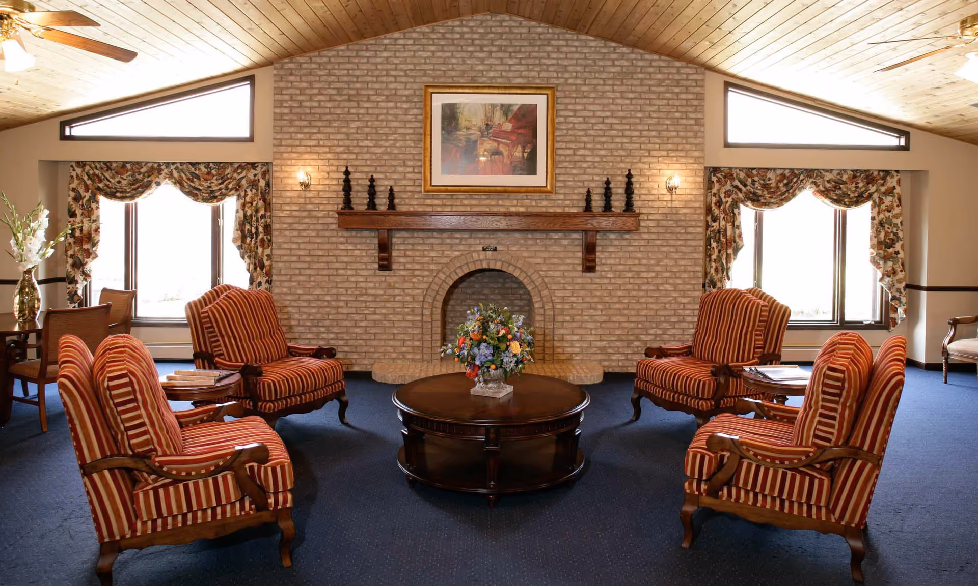 A cozy living room with a high wooden ceiling and a brick fireplace in the center. Four red and beige striped armchairs are arranged around a round wooden coffee table with a floral centerpiece. Two large windows with floral curtains flank the fireplace, allowing natural light to fill the room. There is a framed painting above the fireplace and a ceiling fan with lights.