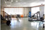 Exercise room with treadmills, stationary bikes, mats, and an exercise ball near large windows with vertical blinds.
