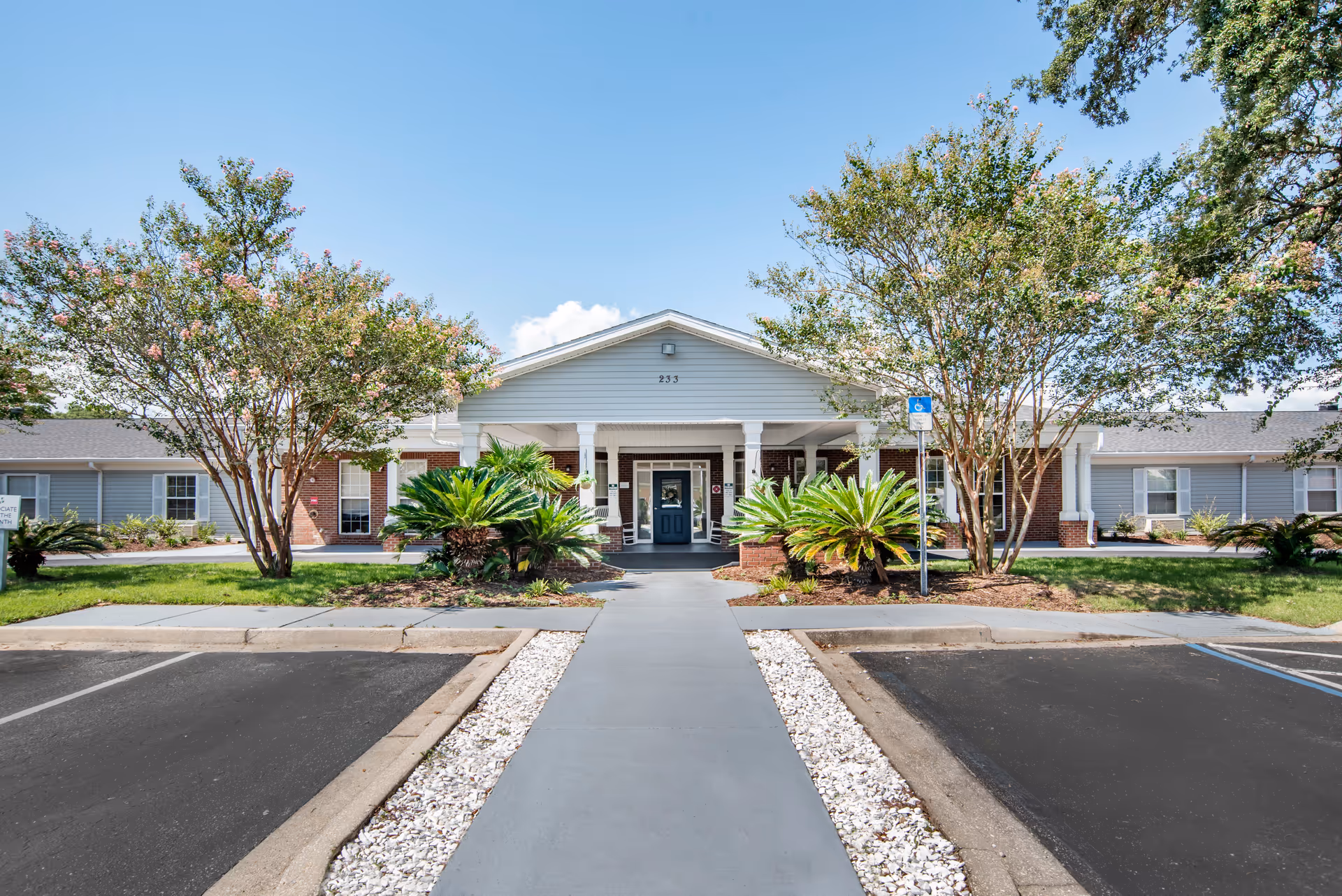 Front entrance of a single-story senior living building with a covered portico, landscaped shrubs and a walkway from the parking lot.
