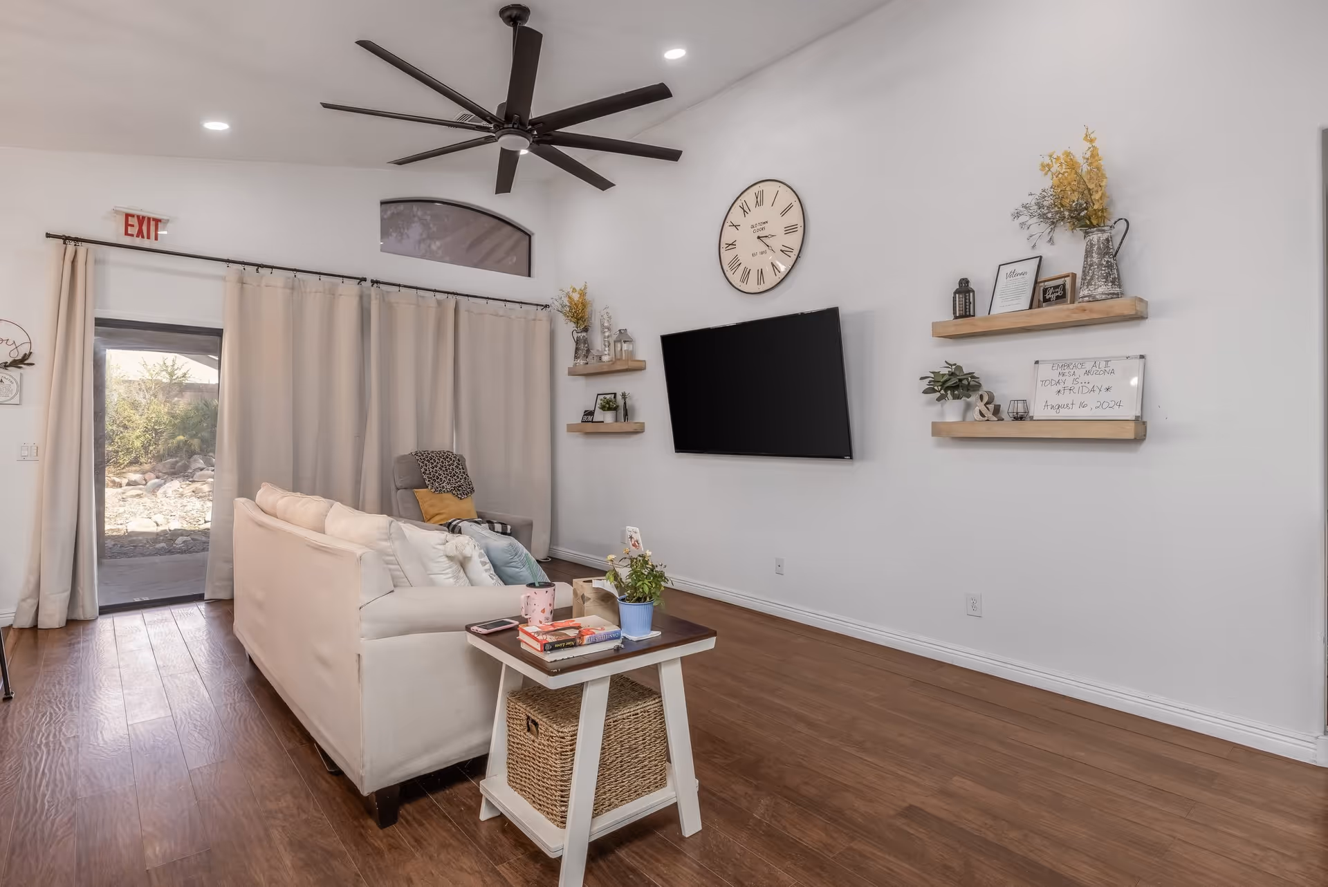 A cozy living room with a beige sofa, a small side table with books and a plant, a large wall-mounted flat screen TV, three wooden floating shelves with decorative items, a large wall clock, and a ceiling fan. The room has wooden flooring and a glass door with beige curtains leading outside.