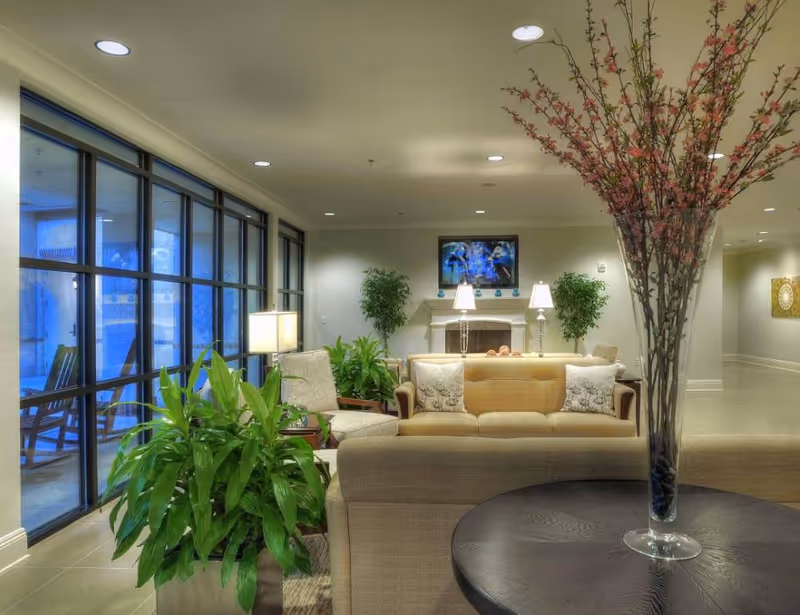 A cozy senior living facility lounge area with beige sofas and armchairs arranged around a fireplace. There are two table lamps on the mantel, green potted plants, and a large vase with tall branches and pink flowers on a round wooden table in the foreground. Large windows line one side of the room, letting in natural light.