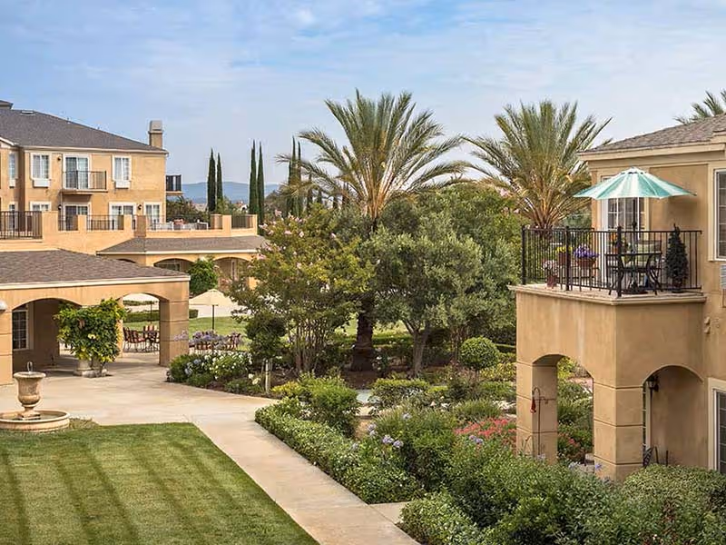 Landscaped courtyard of an apartment-style senior living community with palm trees, walkways, a fountain, and balconies.