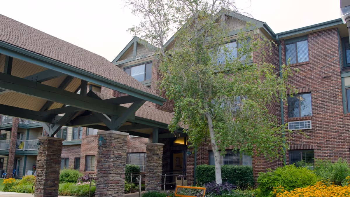 Exterior view of a multi-story brick building with a covered entrance supported by stone pillars. There are windows with blinds, green shrubs, a tree, and yellow flowers in the landscaped area in front of the building.