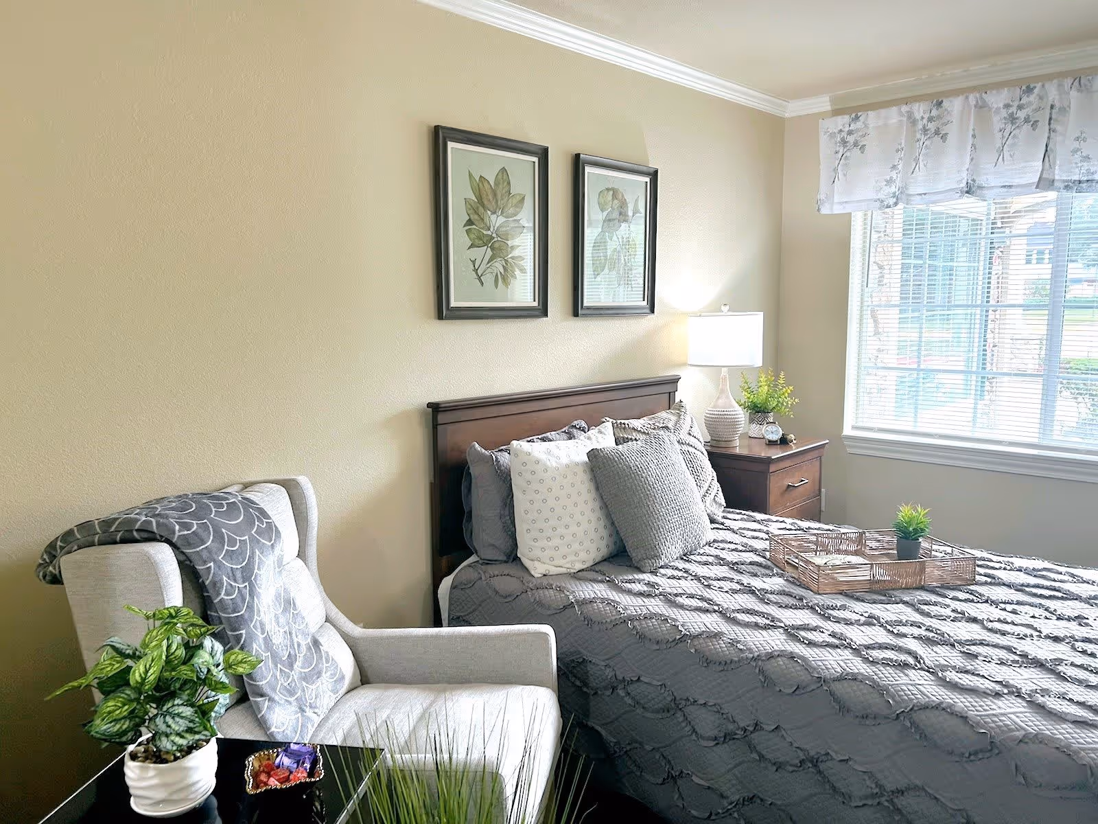 A cozy bedroom with a neatly made bed featuring gray bedding and multiple pillows. Next to the bed is a wooden nightstand with a lamp, a small plant, and an alarm clock. A comfortable armchair with a patterned throw blanket is positioned beside a small table holding a potted plant and a dish with candy. Two framed botanical prints hang on the beige wall above the bed. A large window with floral curtains allows natural light to brighten the room.