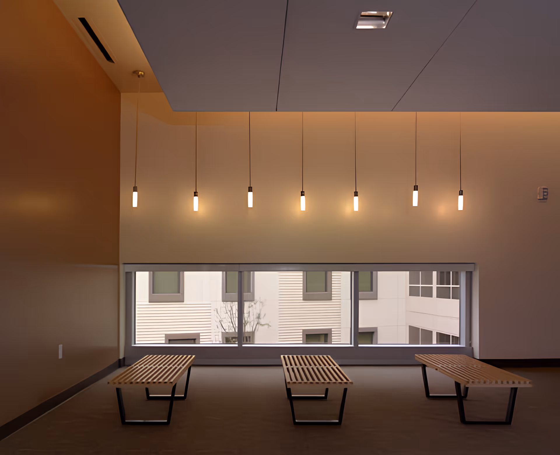 A modern interior space with three wooden benches with black metal legs arranged in front of a large horizontal window. Above the window, seven pendant lights hang from the ceiling, casting a warm glow. The walls are painted in neutral tones, and the ceiling has a recessed lighting fixture.
