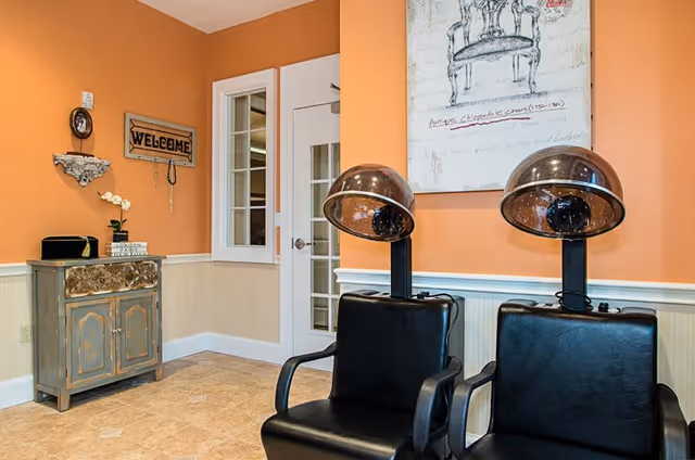 Interior room with two black salon hair dryer chairs, an orange and beige wall, a small vintage cabinet with decorative items, a 'WELCOME' sign on the wall, and a framed artwork above the chairs.