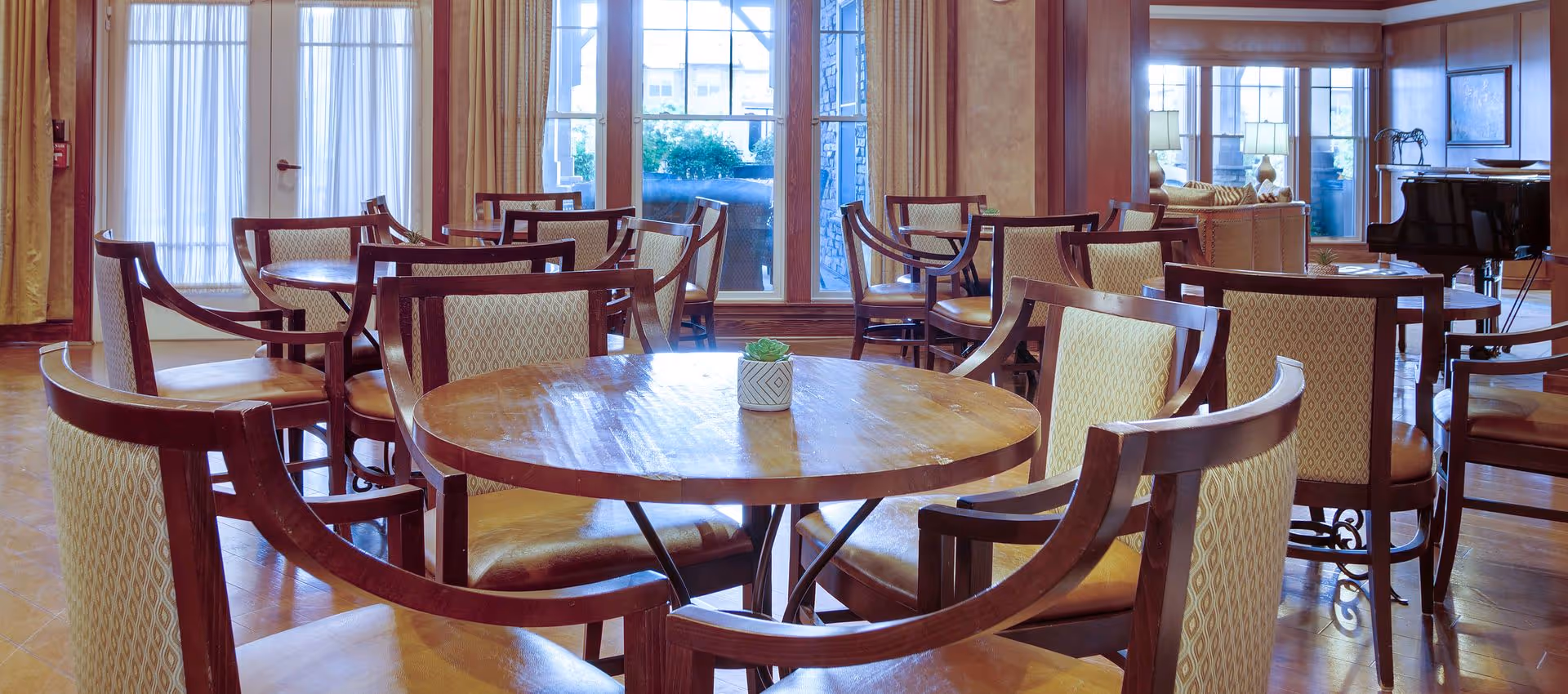 Bright communal dining area with round wooden tables and upholstered chairs by large windows.