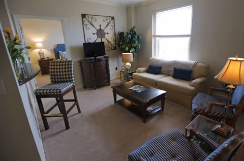 A cozy living room with a beige sofa adorned with two dark blue cushions, two striped armchairs, a wooden coffee table with magazines, a small TV on a dark wooden dresser, a large decorative wall piece, a potted plant, and two table lamps providing warm lighting. A doorway leads to a bedroom with a blue headboard visible.