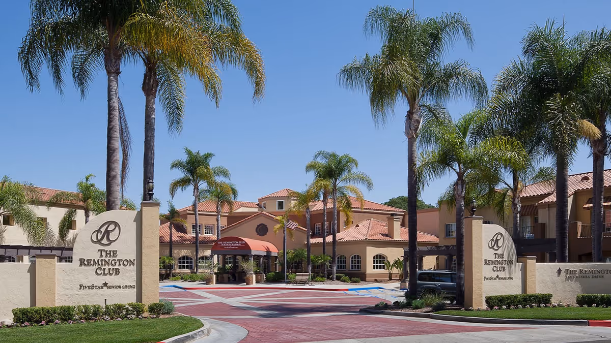 Entrance to The Remington Club senior living facility with palm trees lining the driveway, beige buildings with red tile roofs, and signage displaying the facility's name.