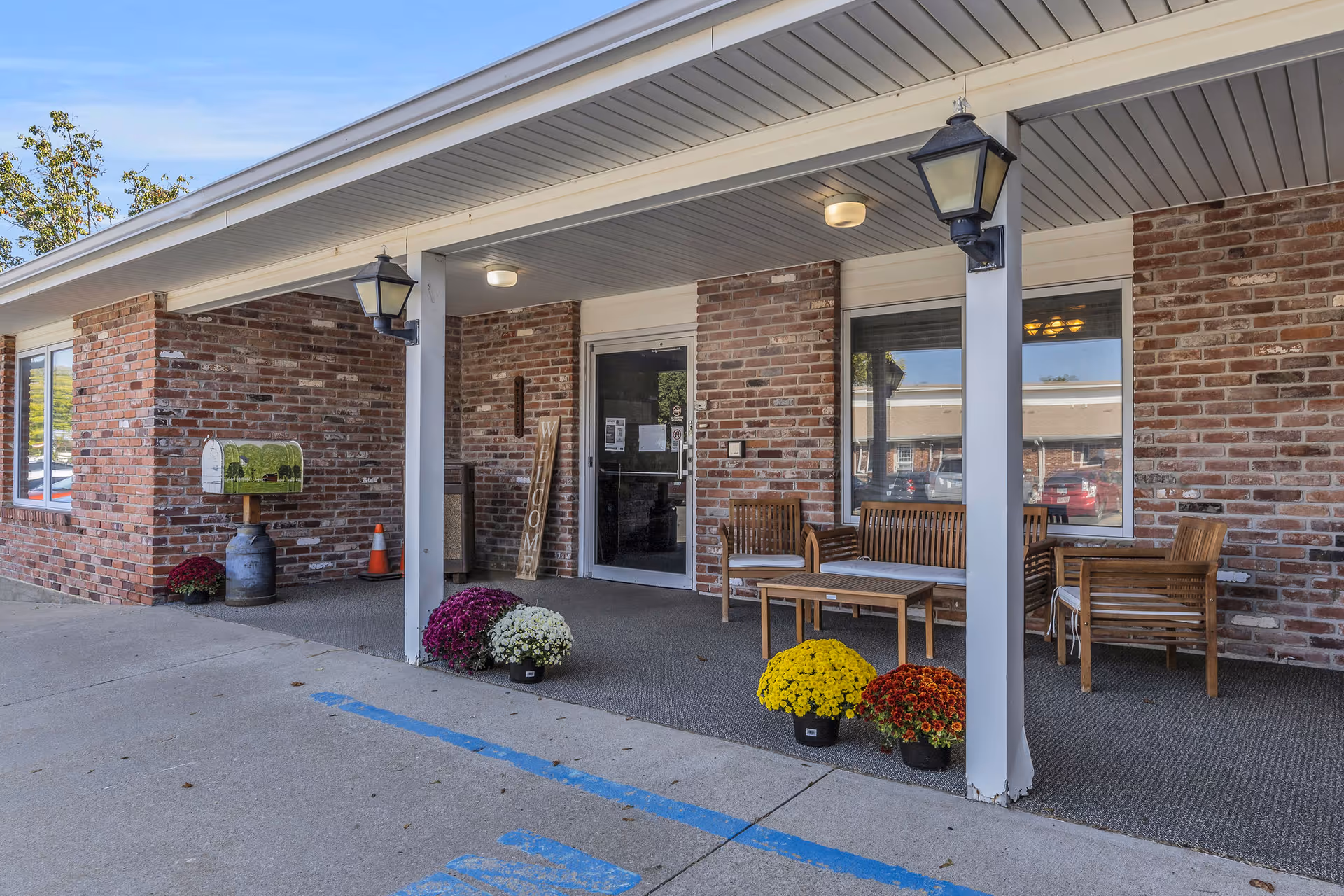 Entrance area of a brick building with a covered porch featuring wooden outdoor furniture including chairs and a bench with a table. There are colorful potted flowers placed on the ground near the entrance. Two lantern-style lights hang from the porch ceiling, and a 'WELCOME' sign is leaning against the wall next to the glass door entrance.
