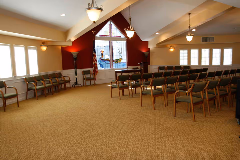 A large room with rows of wooden chairs with green cushions arranged facing a podium and a stained glass window featuring a cross. The room has beige walls with a red accent wall behind the podium, carpeted floor, and several windows letting in natural light. There is an American flag next to the podium.