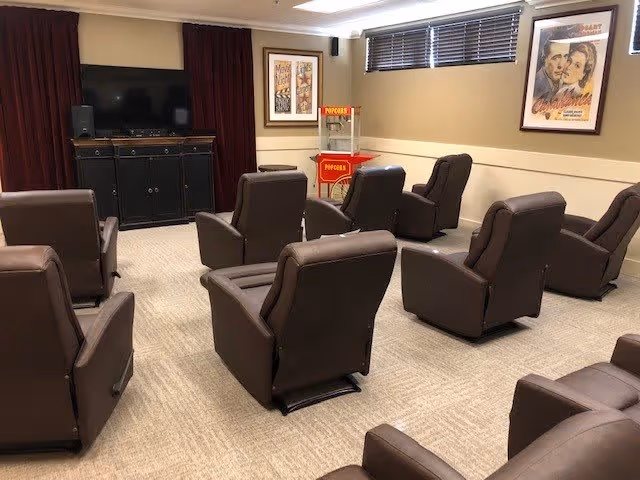 A small movie theater room with eight brown recliner chairs arranged in two rows facing a large flat-screen TV mounted on a black cabinet. There is a popcorn machine in the back corner and framed vintage movie posters on the walls. The room has beige walls and carpet, with a window covered by blinds.