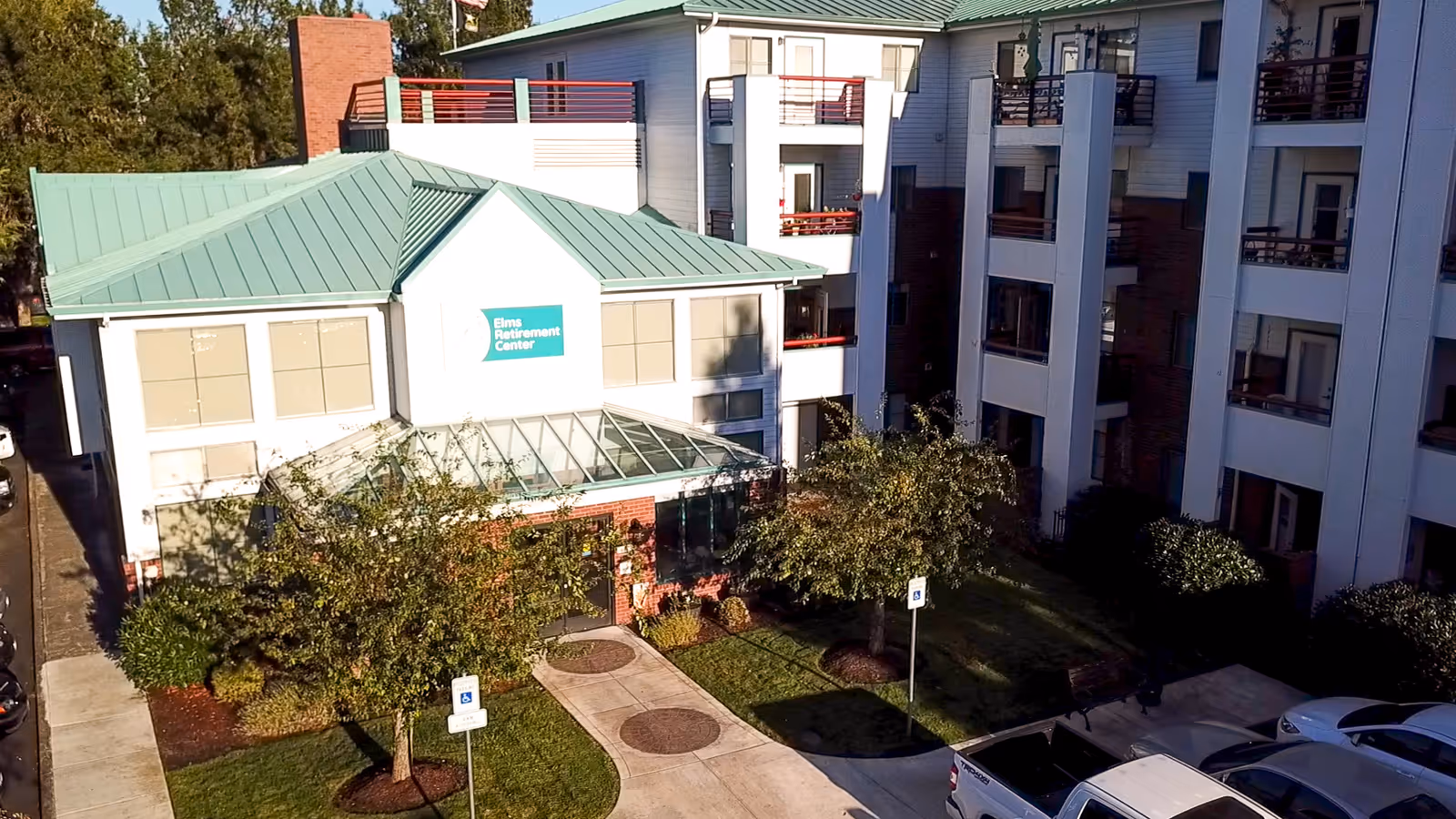 Exterior view of Elms Residential Center, a multi-story building with white walls and green metal roofs. The building has balconies and a glass entrance area surrounded by trees and landscaping. There are parking spaces and a sidewalk leading to the entrance.