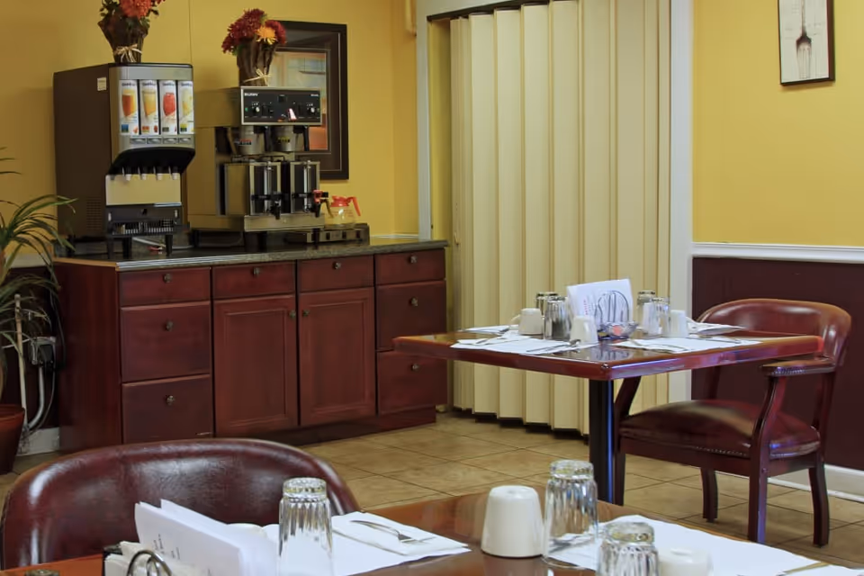 Interior view of a dining area with wooden tables and chairs. Tables are set with upside-down glasses, napkins, and menus. Against the wall, there is a wooden cabinet with a beverage dispenser and a coffee machine on top. The walls are painted yellow with a framed picture and a potted plant nearby.