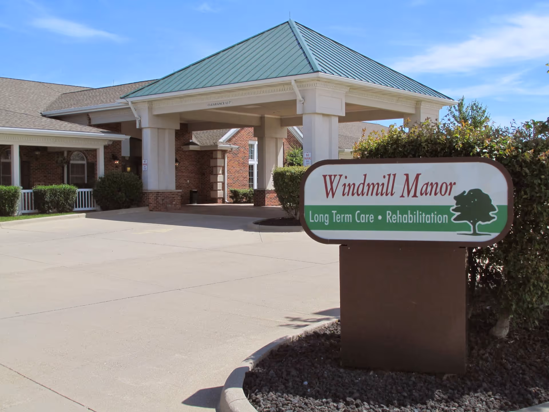 Exterior view of Windmill Manor, a long term care and rehabilitation facility, showing the entrance with a covered drop-off area and a sign with the facility name and services in front of some bushes.