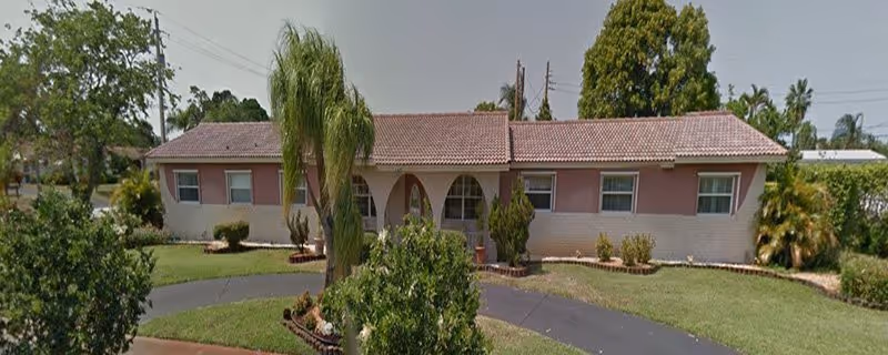 Single-story residential building with a tiled roof, light pink and white exterior walls, arched windows and doorways, surrounded by a well-maintained lawn with palm trees and shrubs. A paved driveway curves in front of the building.