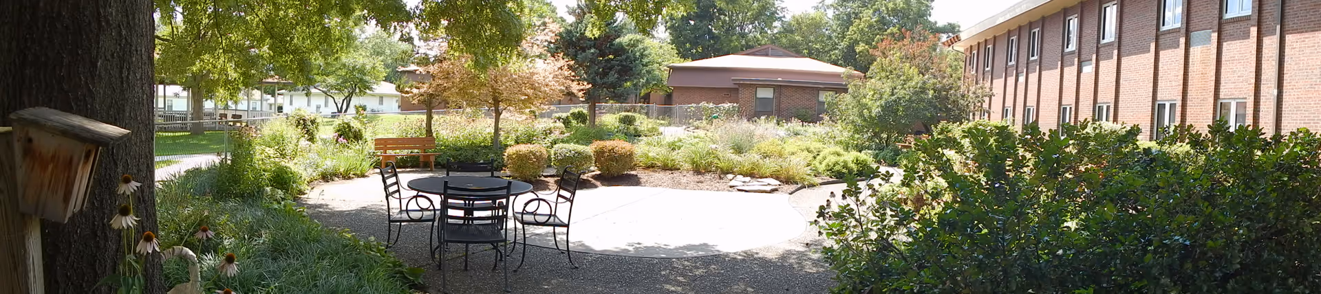 Landscaped courtyard with a round patio table and chairs, benches, garden beds, and the brick exterior of a senior living building.