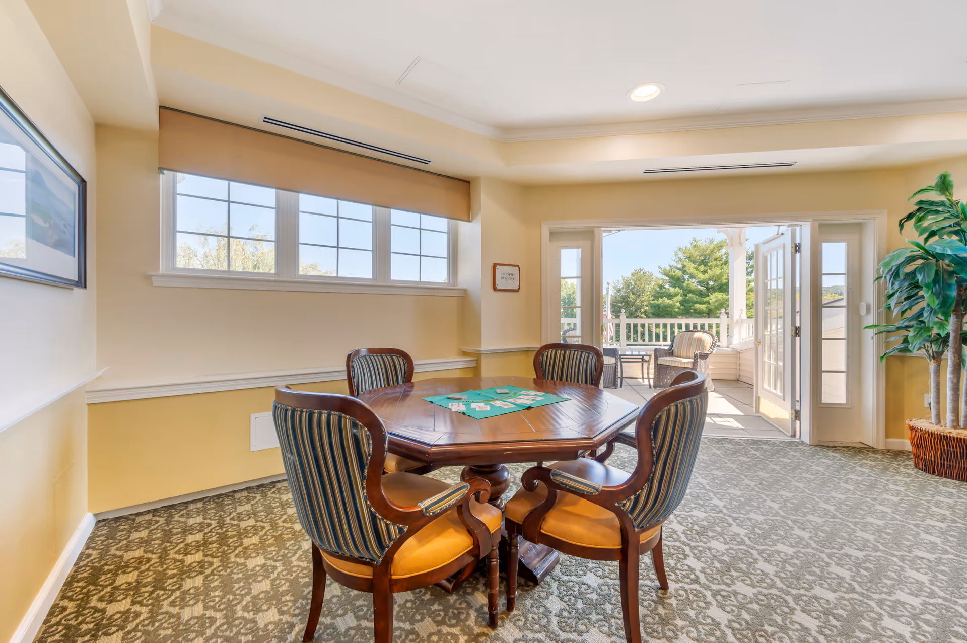 A bright room with a round wooden table and four upholstered chairs arranged around it. Playing cards are spread out on the table. The room has patterned carpet, cream-colored walls, and a large window letting in natural light. French doors open to an outdoor balcony with wicker chairs and a table, surrounded by greenery.