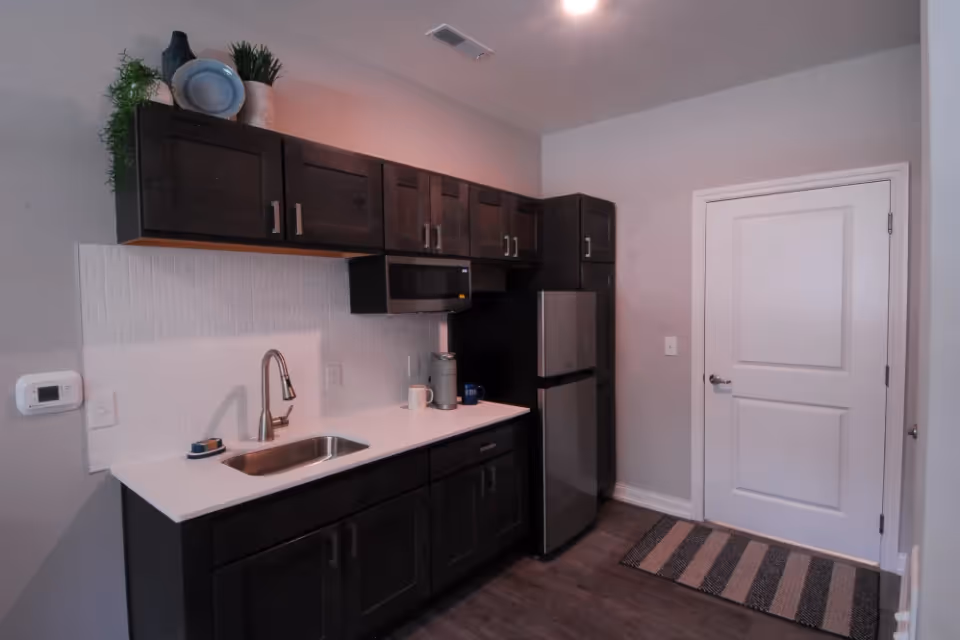 A modern kitchen area with dark wood cabinets, a stainless steel sink, microwave, and refrigerator. The countertop is white with a few items including a coffee mug and a small container. There is a white door on the right side and a striped rug on the floor in front of it. The walls are light-colored and the floor is a dark wood finish.