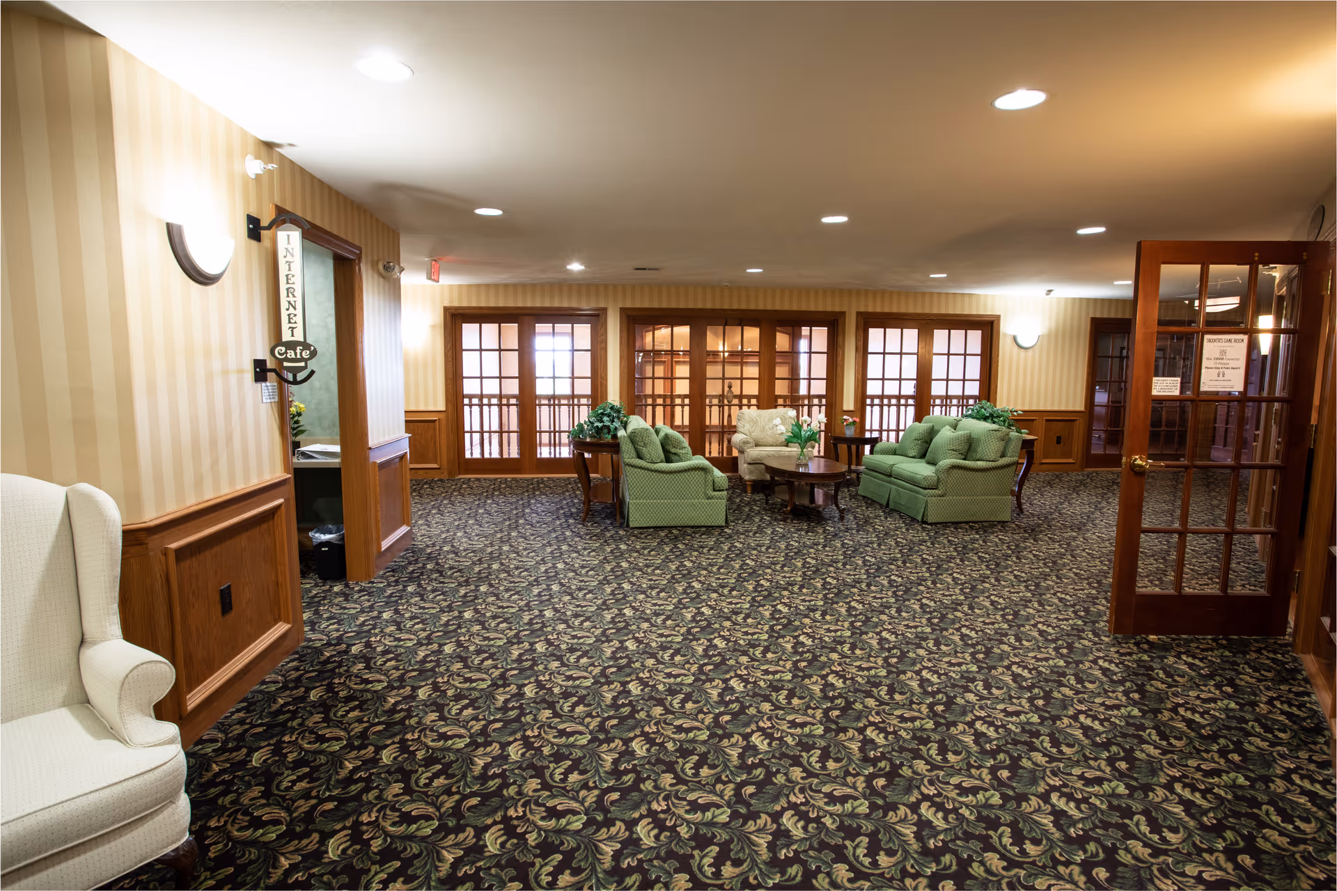 A spacious senior community lounge area with patterned carpet, beige striped wallpaper, and wooden paneling. The room features green upholstered sofas and armchairs arranged around a wooden coffee table with plants on it. There are multiple wooden-framed glass doors along the back wall, and a sign on the left points to an Internet Cafe. The lighting is warm with ceiling lights and wall sconces.