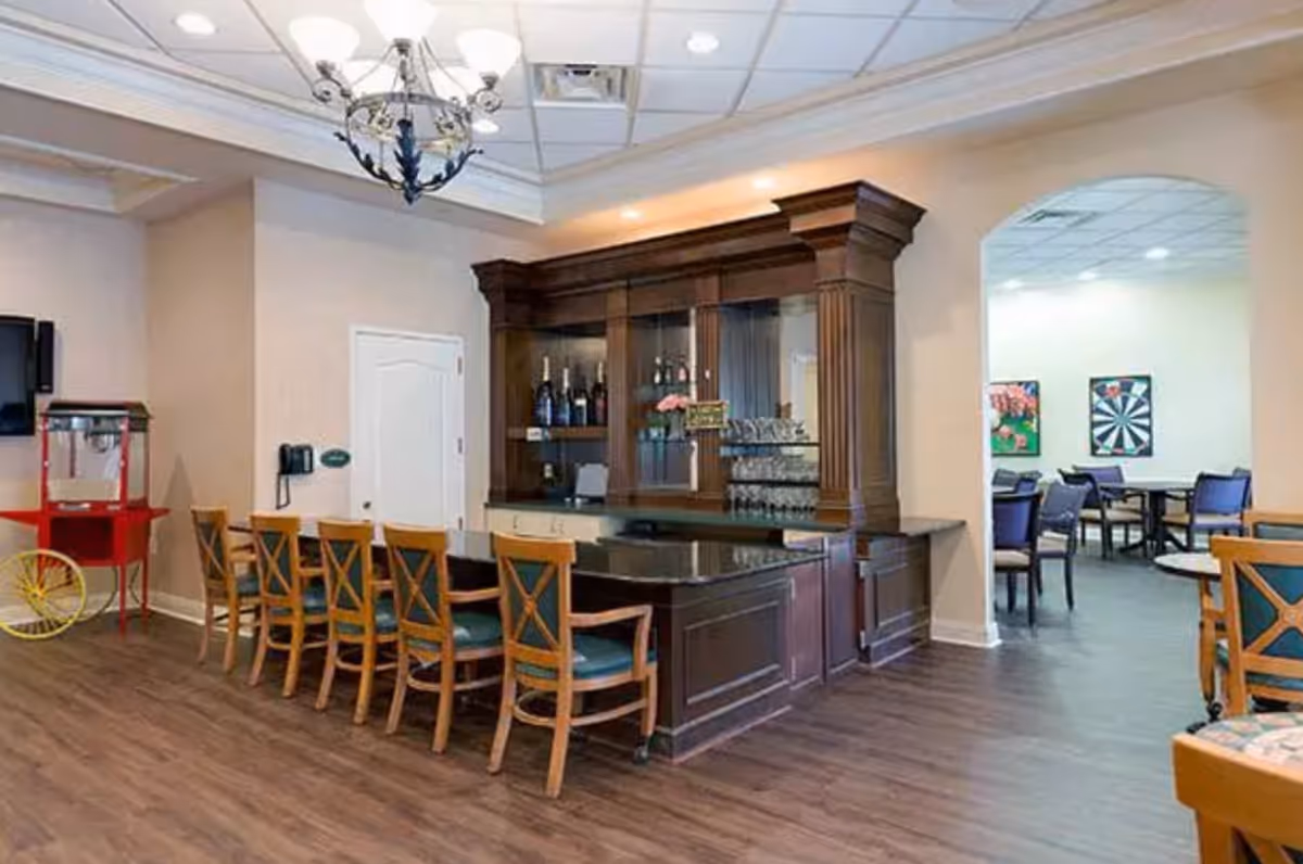 Interior view of a senior living facility featuring a wooden bar counter with several chairs lined up in front. Behind the bar, there are shelves with bottles and glassware. To the left, there is a red popcorn machine and a wall-mounted telephone. In the background, through an archway, there is a room with tables and chairs, and colorful artwork on the walls. The floor is wooden, and the ceiling has recessed lighting and a chandelier above the bar area.