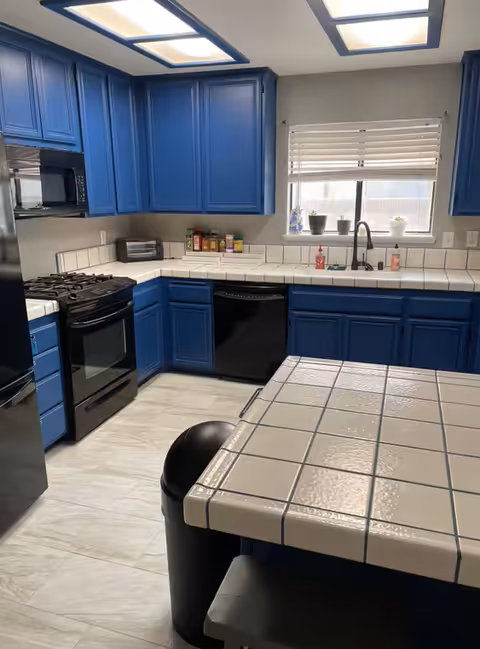 A modern kitchen with blue cabinets, white tiled countertops, and a tiled island. The kitchen features a black stove, microwave, dishwasher, and refrigerator. There is a window above the sink with white blinds and small potted plants on the windowsill. The floor has light-colored tiles and there is a black trash can next to the island.