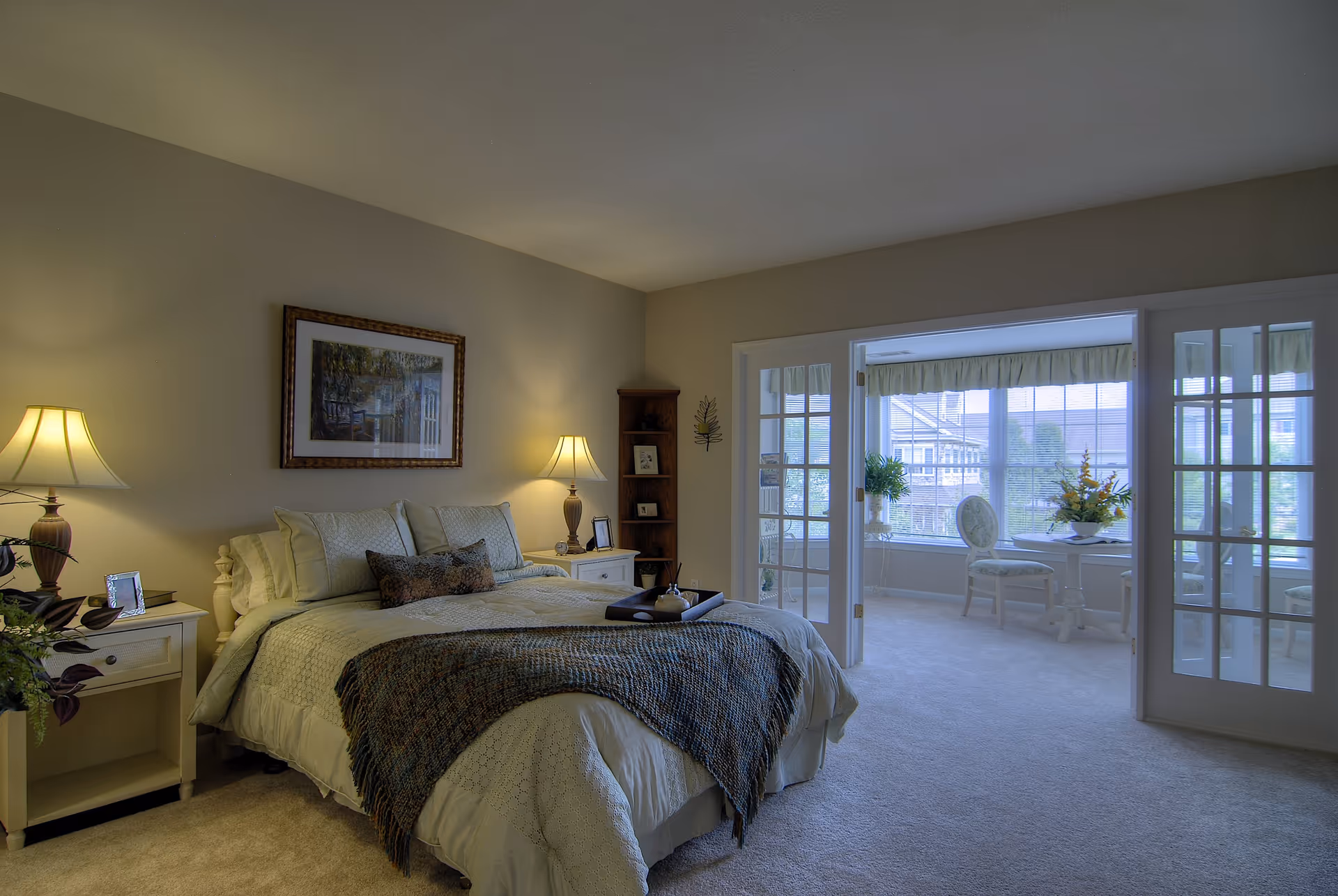 A cozy bedroom with a neatly made bed featuring multiple pillows and a textured throw blanket. Two bedside tables with lamps flank the bed. A framed picture hangs above the bed. French doors open to a bright sunroom with large windows, a small round table, and two chairs, decorated with plants and flowers.