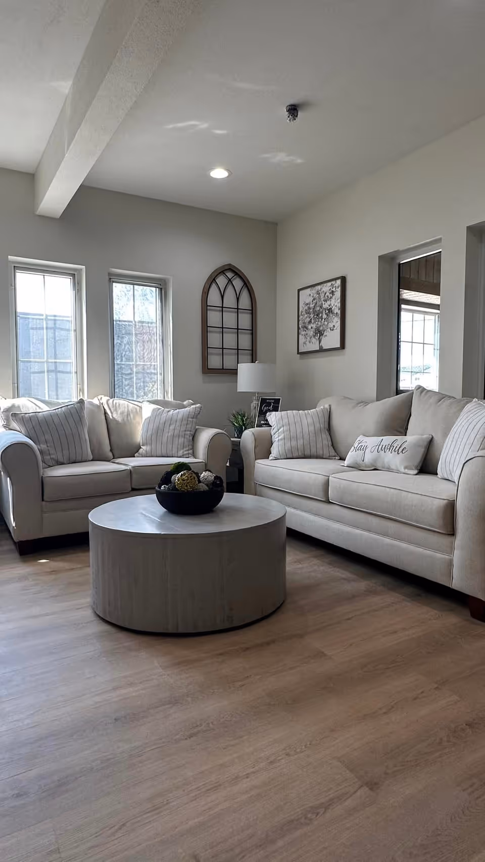 A cozy living room with two beige sofas adorned with striped and plain cushions, a round wooden coffee table with a decorative bowl on top, light wood flooring, and wall decor including a framed picture and an arched window frame. Natural light enters through three tall windows.
