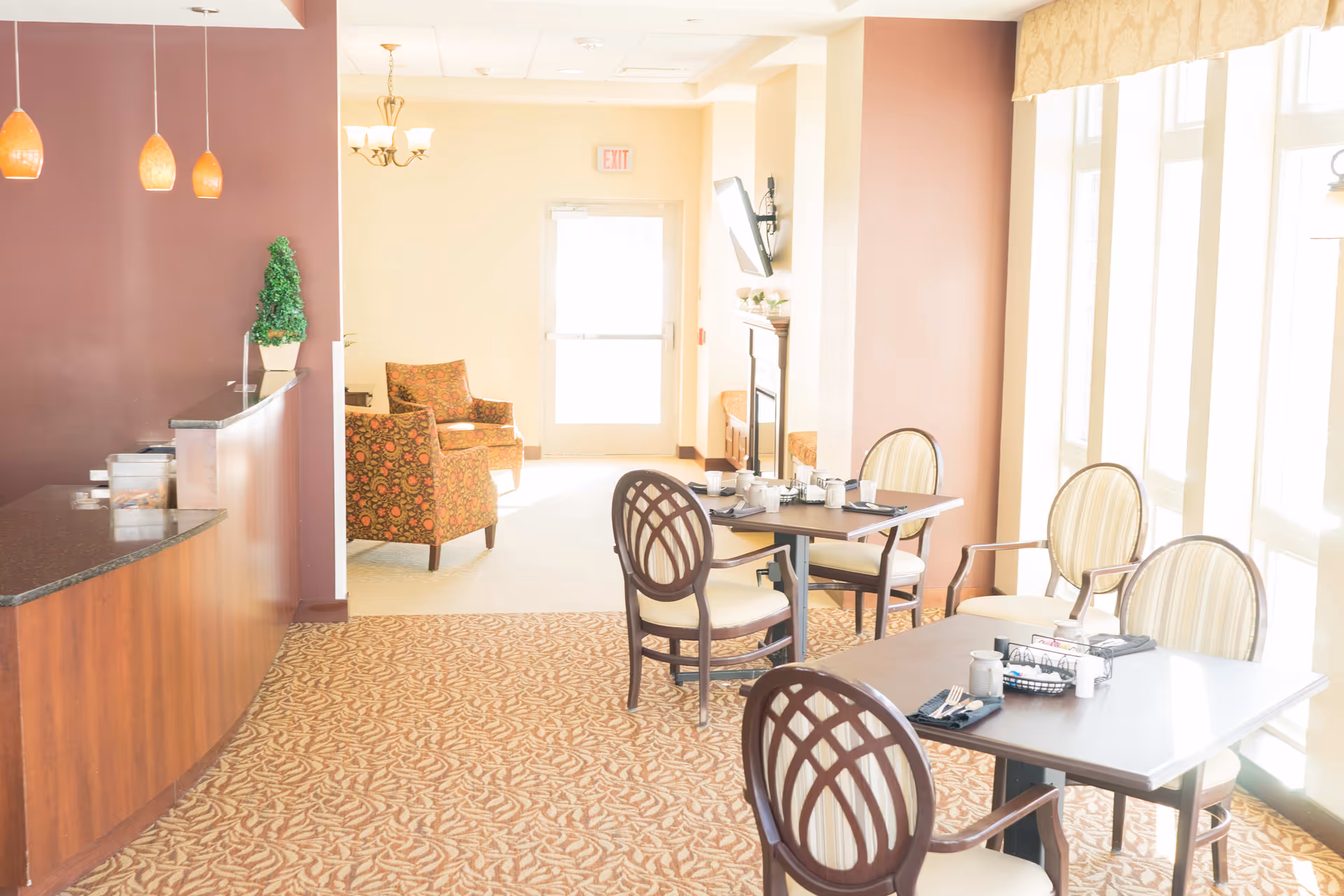 A bright dining area in a senior living facility with tables and chairs arranged for meals. The room features large windows letting in natural light, a patterned carpet, a small counter with pendant lights, and a seating area with two upholstered armchairs near a door.