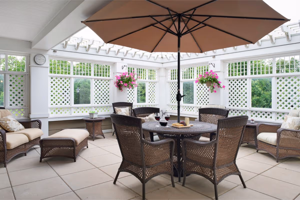 A bright enclosed patio area with a round table and six wicker chairs under a large brown umbrella. The table has a wine decanter, two glasses of red wine, a plate with cheese and crackers, and grapes. There are two hanging flower baskets with pink flowers on the white lattice walls, and cushioned wicker armchairs and an ottoman are arranged around the space. A clock is mounted on one wall, and greenery is visible through the windows.