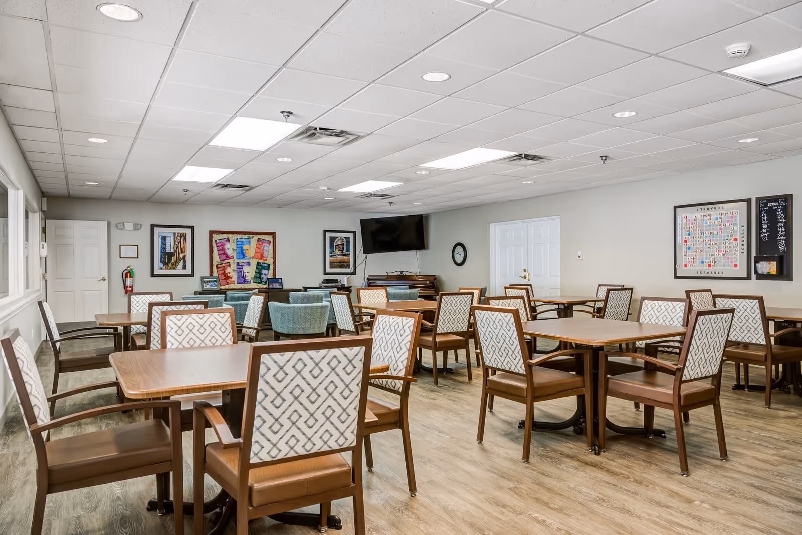 A senior living facility common room with multiple wooden tables and chairs arranged neatly. The chairs have patterned upholstery. The room has a light-colored wood floor, white walls, and a drop ceiling with recessed lighting. On the walls are framed pictures, a large Scrabble board with scores, a wall clock, and a mounted flat-screen TV above a piano.
