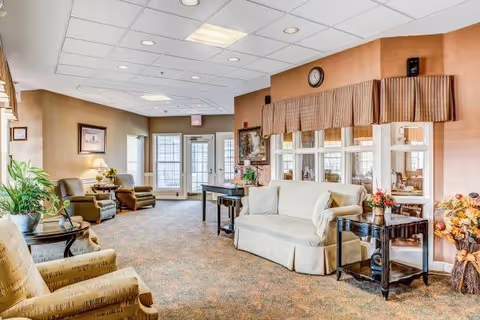A bright and spacious senior living facility common area with beige and brown tones. The room features multiple comfortable armchairs and a sofa arranged around small tables with plants and flowers. Large windows and glass doors let in natural light, and a clock is mounted on the wall above a window with striped valances. The carpet has a patterned design, and the ceiling has recessed lighting.
