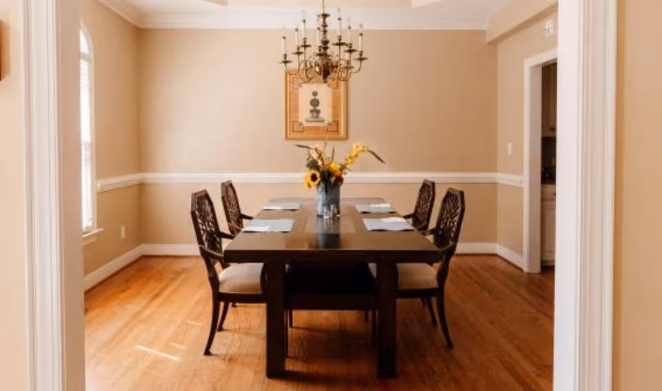 A dining room with a dark wooden table set for six with placemats and a vase of flowers in the center. The room has beige walls, wooden flooring, a chandelier hanging above the table, and a framed picture on the wall.