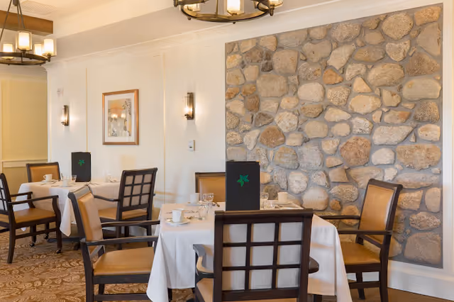 Dining room with tables covered in white tablecloths, set with cups, glasses, and menus. The room features wooden chairs with tan cushions, a stone accent wall, framed artwork, and warm lighting fixtures on the ceiling and walls.
