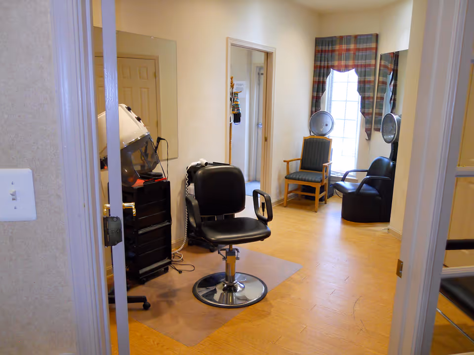 Interior view of a senior care community hair salon area with a black salon chair in the center, a hair dryer station with two hooded dryers near a window with plaid curtains, and a wooden chair with green upholstery. The room has light-colored walls and wood flooring.