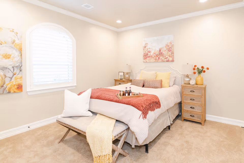 A bright and cozy bedroom with a neatly made bed featuring white and yellow pillows, a red throw blanket, and a tray with drinks. There is a cushioned bench at the foot of the bed with a white pillow and a yellow throw. The room has beige walls, a window with white shutters, two wooden nightstands with lamps, and floral artwork on the walls.