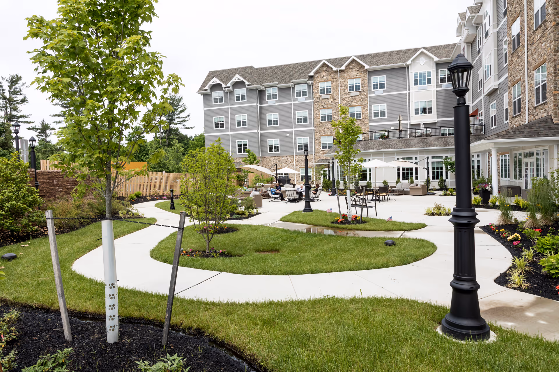 Outdoor courtyard area of a senior living facility with a winding concrete pathway, green grass, young trees, flower beds, black lamp posts, and seating areas with umbrellas. The multi-story building in the background features gray siding and stone accents.