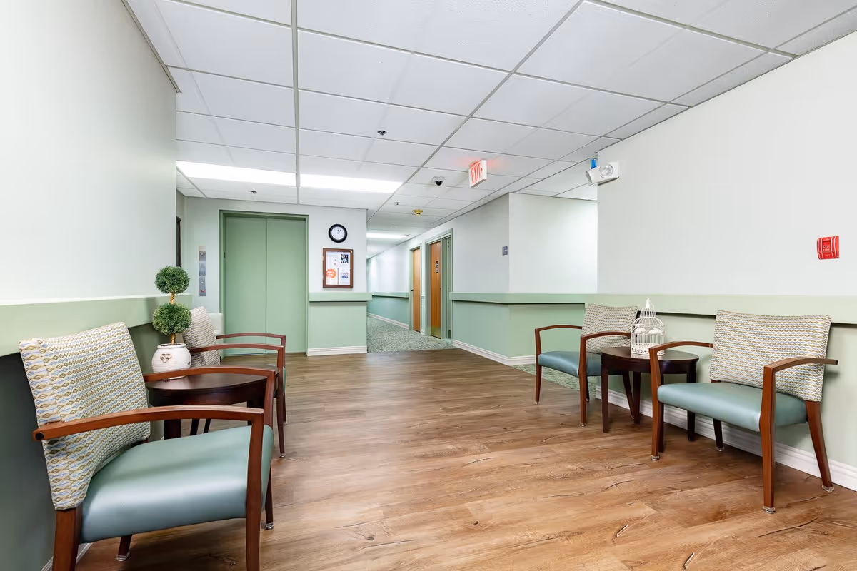 A clean and well-lit hallway in a senior living facility with light green walls and wood flooring. There are four cushioned chairs with wooden arms and patterned pillows arranged in pairs on either side of the hallway, each pair accompanied by a small round wooden table. Decorative items including a small potted plant and a white birdcage are placed on the tables. At the end of the hallway, there is a green elevator door, a clock, and a bulletin board on the wall. The ceiling has white tiles with fluorescent lighting and an exit sign is visible.