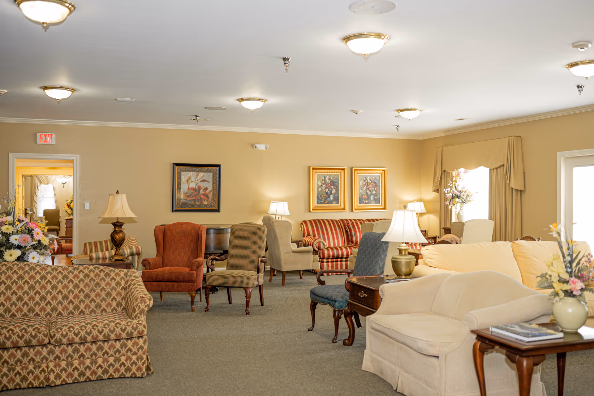 A spacious living room area in Magnolia Estates with various upholstered chairs and sofas arranged around wooden side tables with lamps. The walls are beige with framed floral artwork, and there are floral arrangements on tables. The room is well-lit with ceiling lights and natural light from a window with beige curtains.