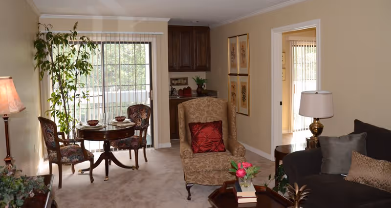 A cozy living room and dining area in a senior living facility with beige walls and carpet. The living room features a patterned armchair with a red pillow, a dark sofa with gray and beige pillows, a wooden coffee table with books and a flower vase, and a side table with a lamp. The dining area has a round wooden table set with two bowls and two floral upholstered chairs. A tall potted plant is near the sliding glass door with vertical blinds, leading to an outdoor space. There are framed botanical prints on the wall and a small kitchenette area with dark wooden cabinets in the background.