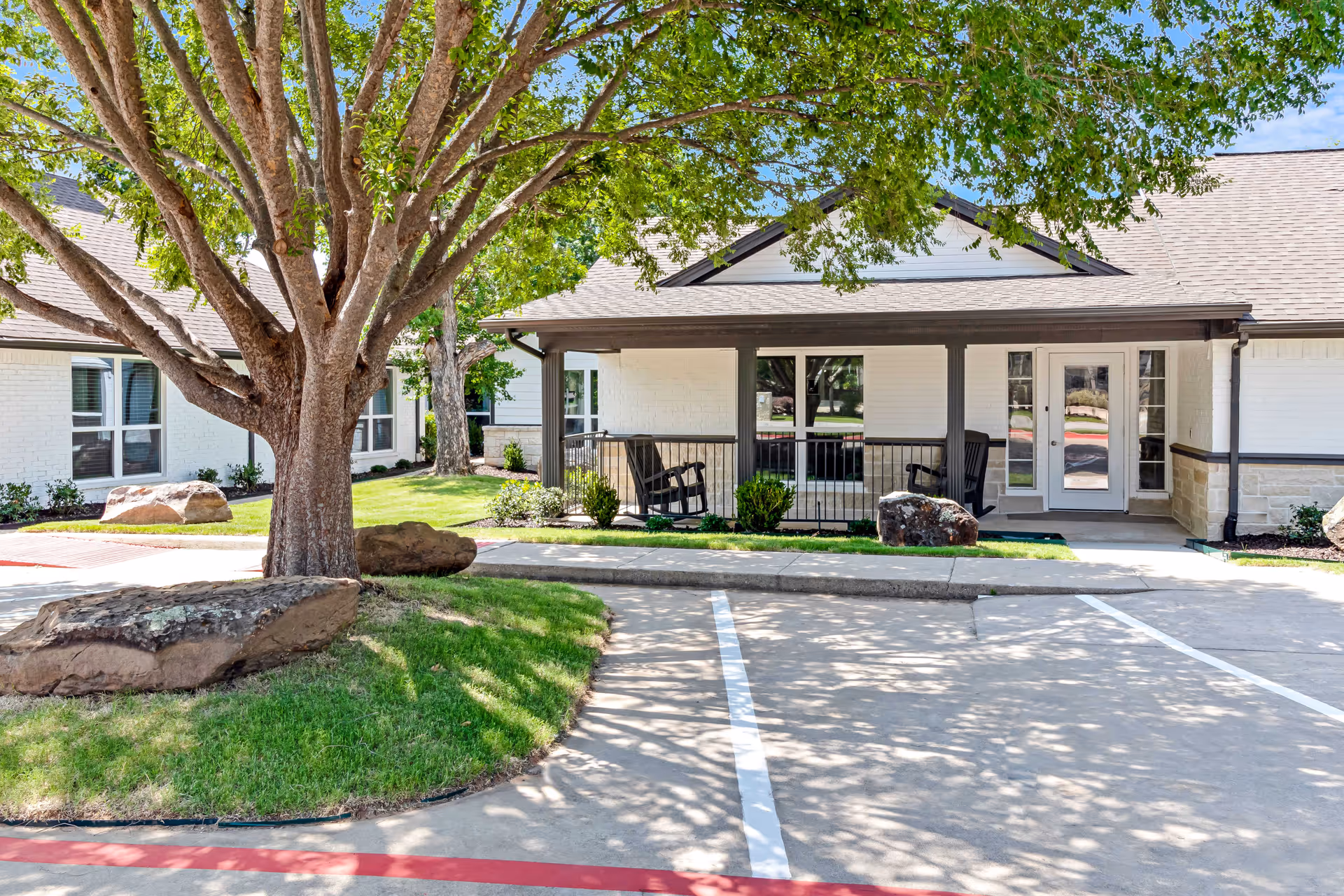 Exterior view of a single-story building with white brick walls and a covered porch area featuring two rocking chairs. There is a large tree with green leaves providing shade over a grassy area with large rocks. The foreground shows an empty parking area with marked parking spaces and a red curb.