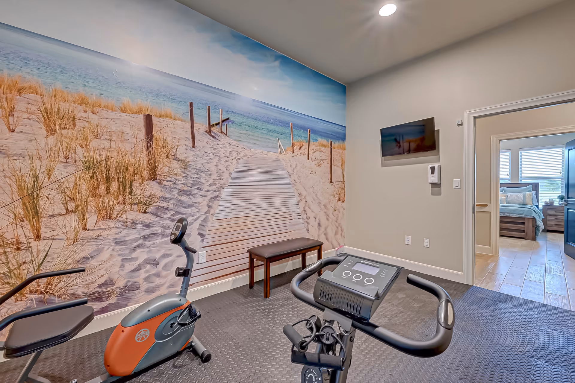 Exercise room with stationary bikes, a bench and wall-mounted TV, a large beach mural on one wall, and a doorway leading to a bedroom.