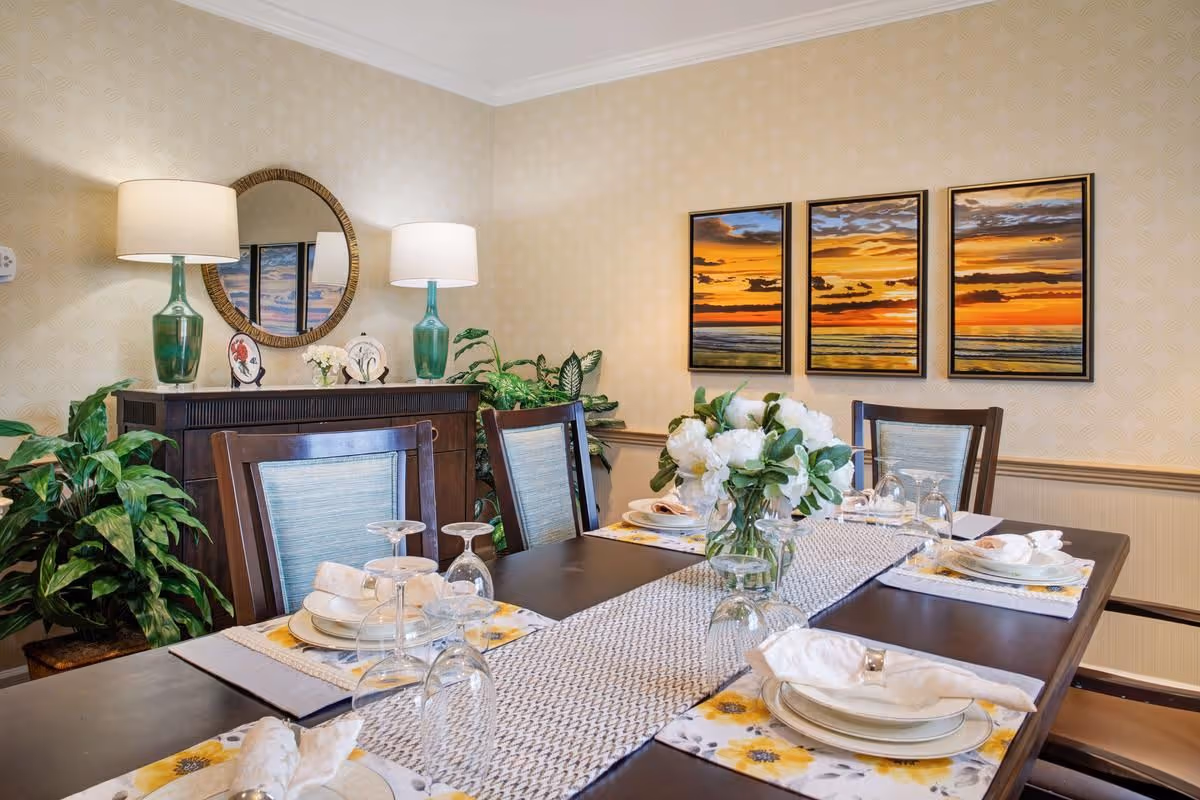 A dining room with a dark wooden table set for six people, featuring white plates, napkins, and upside-down wine glasses. The table has a patterned runner and a vase with white flowers as a centerpiece. Behind the table is a wooden sideboard with two green glass lamps and decorative plates, a round mirror on the wall, and three framed sunset paintings. There are also green plants placed around the room.
